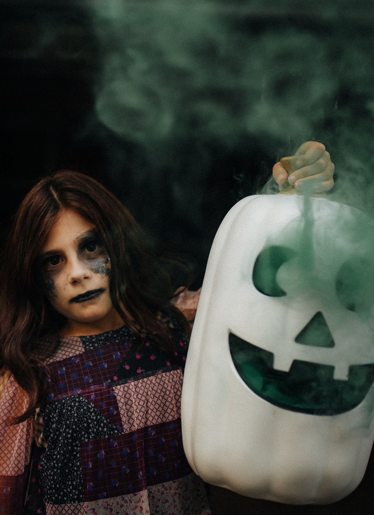 Halloween photo of a girl dressed in halloween make-up holding a white pumpkin with green smoke coming from the eyes